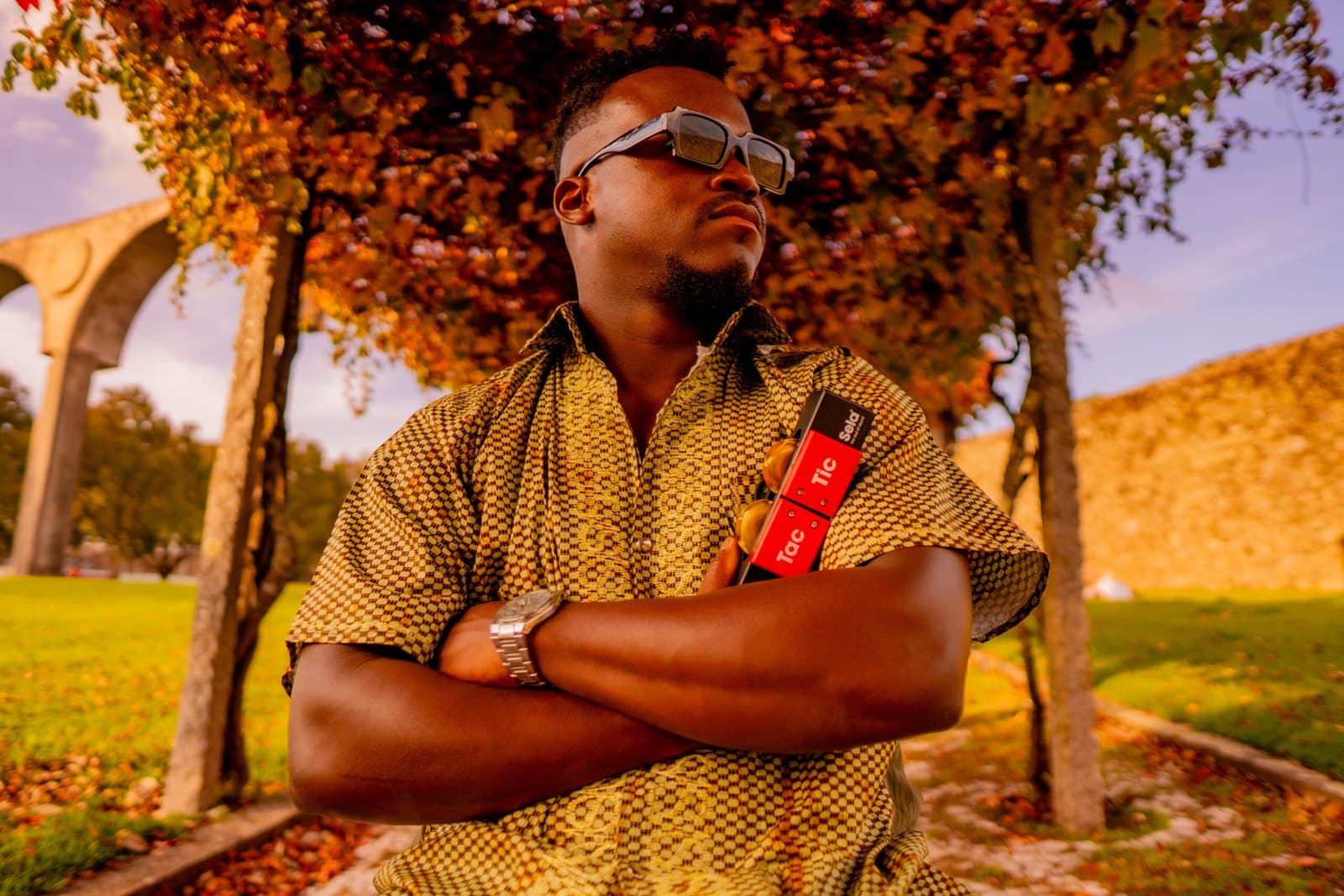 Ivirson de braços cruzados com um shaker Sela Tic Tac, num parque com um aqueduto ao fundo