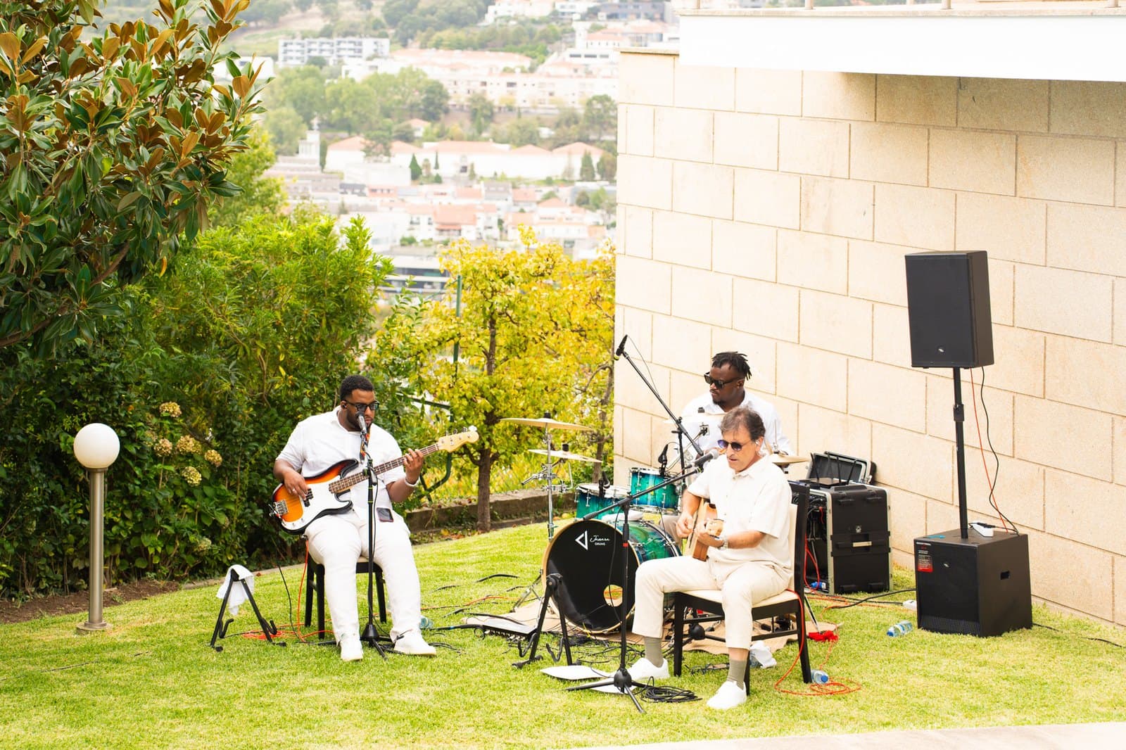 A banda a tocar num jardim relvado: baixo, bateria e guitarra, com colunas de som e a cidade ao longe