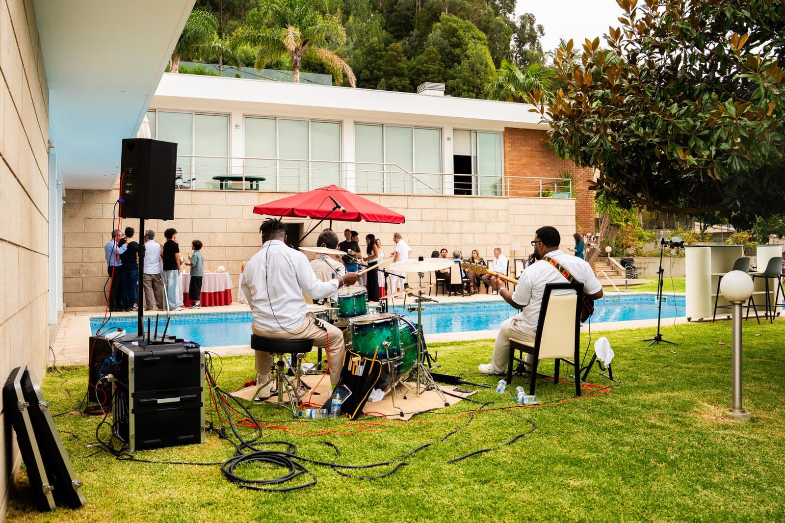 O grupo a tocar junto a uma piscina, com convidados à volta das mesas e um guarda-sol vermelho